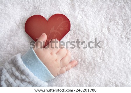 Red heart shaped card in baby hand on white soft blanket background