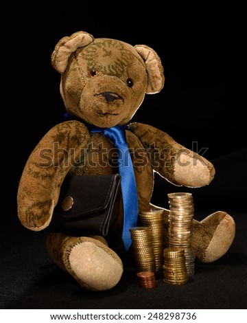 Teddy as a businessman, who proudly presents its stacked money. He has a black briefcase under his arm and stacked coins in front of him. Studio shot isolated against a black background shown.