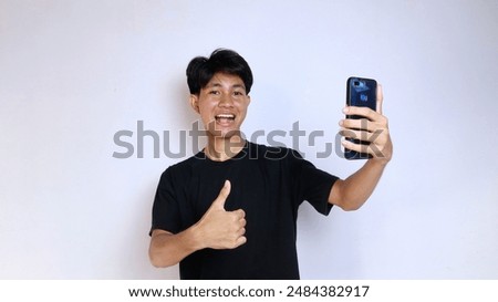 Happy young Asian man wearing black shirt gesturing okay while holding smartphone on isolated white background