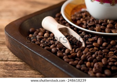 Picture of coffee grains on tray from dark wood with wooden spoon and cup on plate with gold and red color everything placed on old wood.