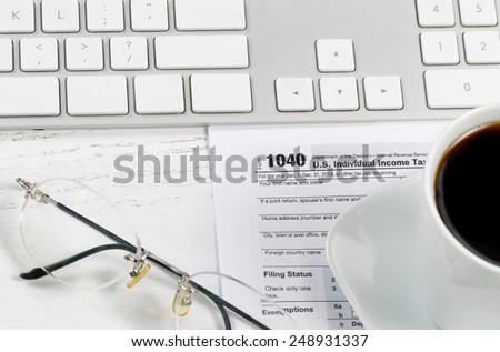 Close up of tax form, reading glasses, computer keyboard and coffee on white desk. Focus on Tax form 1040. 