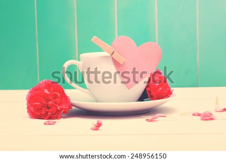 Coffee cup with a pink heart tag and carnations on a teal colored wood background