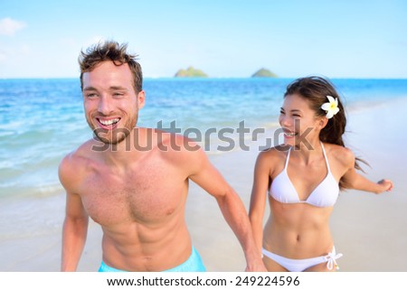 Happy couple having fun on beach vacation during summer holiday. Multiracial fit couple running together holding hands laughing in the sun. Young adults in shape carefree feeling good in their body.