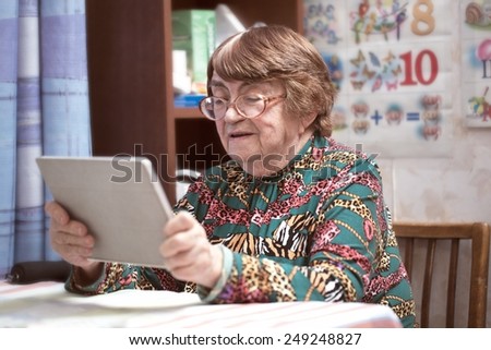 Senior woman in glasses sitting at the table at home and watching photos or video on tablet computer
