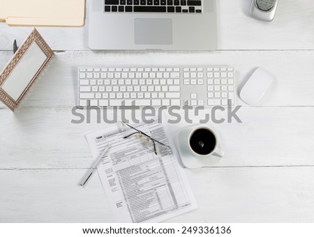 Top view angle of office desktop consisting of laptop, keyboard, pens, mouse, picture frame, phone, coffee, reading glasses, tax forms and work folders on white desk.