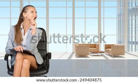 Businesswoman in headset holding clipboard and pen, sitting on office chair in vast white interior with transparent wall, smiling