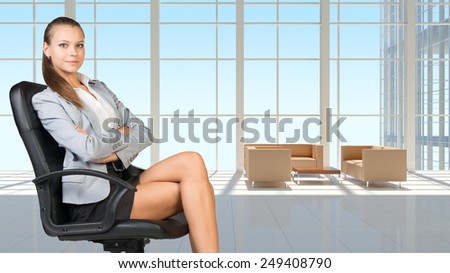 Businesswoman in headset sitting on office chair with her arms crossed, in vast white interior with transparent wall, looking at camera, smiling