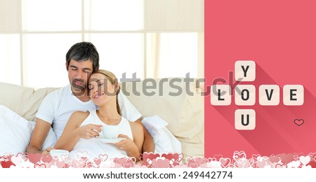 Positive couple drinking coffee lying in the bed against love you tiles