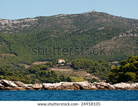 This is a picture of a church on a hill; surronded by sea; trees and mountains and visible sky-strip