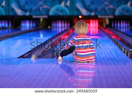 Adorable little child, blonde toddler girl, sitting on the floor at bowling club waiting for a strike - weekend family leisure concept
