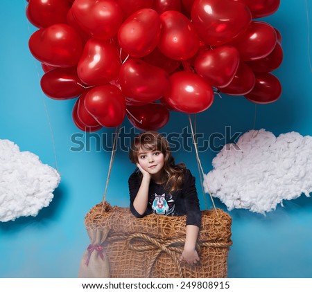 Small brunette girl with beautiful face on a board of aerostat with a lot of red balloons having heart form on top flying in happy mood under bright blue sky with clouds and wind playing with her hair