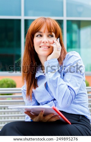 Photo of caucasian businesswoman sitting on a metal bench with charts in her hands and phoning