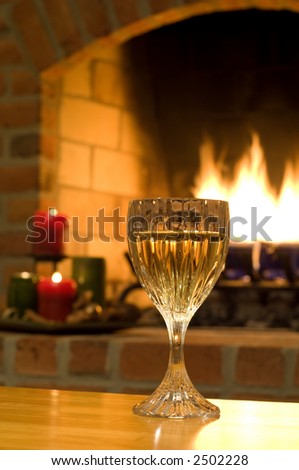 A glass of white wine is highlighted by the flames of a warming fire