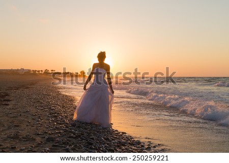 Barefoot young bride in a white wedding dress enjoys a lonesome walk on a wet sandy beach in a late summer hazy day, at dusk.