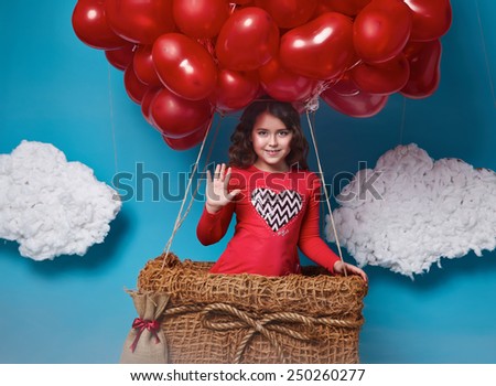 Small cute girl with beautiful face on a board with a lot of red balloons having heart form on  top flying in happy mood under bright blue sky with clouds and wind playing with her hair Valentines day