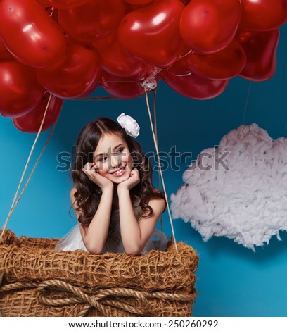 Small cute girl with beautiful face on a board with a lot of red balloons having heart form on  top flying in happy mood under bright blue sky with clouds and wind playing with her hair Valentines day