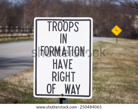 A close up of a metal sign that says Troops In Formation Have Right Of Way along the side of a road.