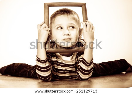Portrait of little funny blonde boy child holding photo frame framing his face looking into the corner studio shot