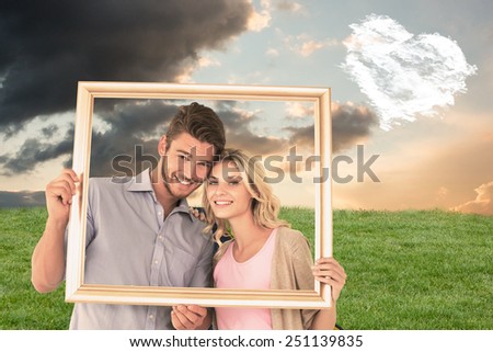 Attractive young couple holding picture frame against cloud heart