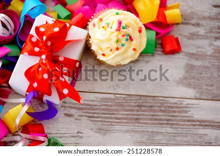 Celebrating a special day. Top view image of multicolored confetti as a frame for a gift box with red ribbon and a cupcake on wooden table with copy space