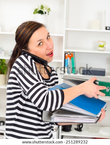 young businesswoman taking a mobile call standing balancing three large office binders on her arm