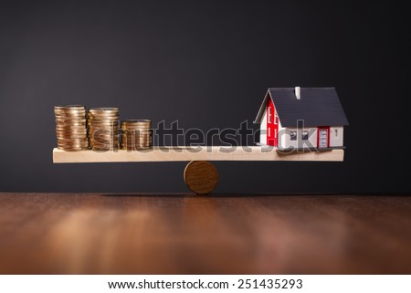 Seesaw with a house on one side and stacks of coins on the other side. Royalty-Free Stock Photo #251435293