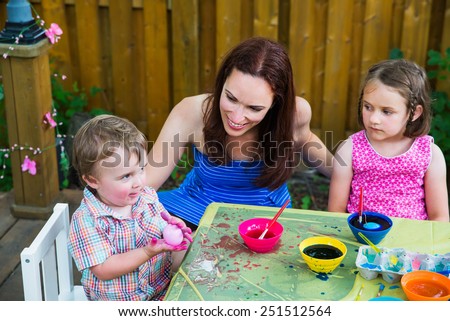 Family picture of a mother smiling at her happy boy holds up a freshly color dyed pink Easter egg outside during the spring season in a garden setting.  A girl watches.  Part of a series.   