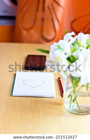 Office composition for St. Valentines Day: bouquet of white freesia flowers, smartphone, pen and notebook with painted heart. Selective focus. Blurred multicolored background. Vertical image
