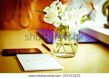 Office composition for St. Valentines Day: bouquet of white freesia flowers, smartphone, pen and notebook with painted heart. Selective focus. Blurred multicolored background. Vintage image