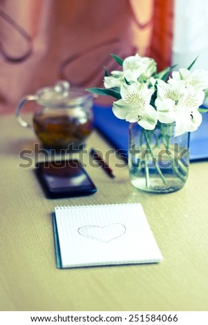 Spring composition for St. Valentines Day: bouquet of white freesia flowers, smartphone, pen and notebook with painted heart. Selective focus. Blurred multicolored background. Vertical vintage image