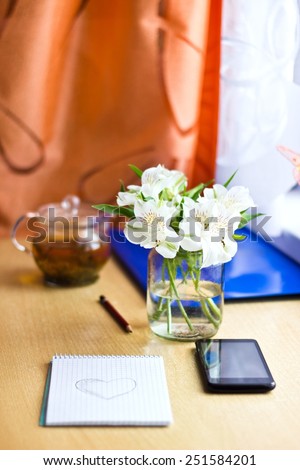 Spring composition for St. Valentines Day: bouquet of white freesia flowers, smartphone, pen and notebook with painted heart. Selective focus. Blurred multicolored background. Vertical image
