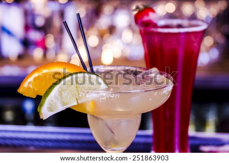Close up of fresh lime margarita with ice cubes in margarita glass sitting on bar top garnished with orange and lime wedges and strawberry vodka drink in background