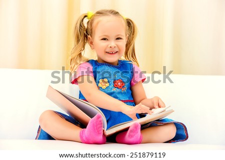 Pretty little girl sitting on a sofa and looking at a children's picture book. Happy childhood.