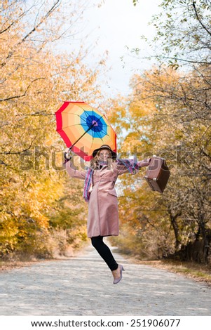 Young lady with rainbow colorful umbrella and suitcase jumping and looking at camera over autumn road