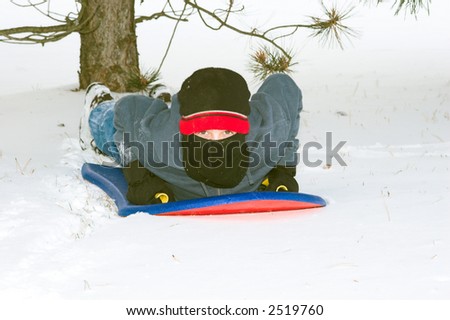 Color DSLR picture of a young boy wearing a mask riding a sleigh in the winter snow. Pine tree in background.  Horizontal orientation with copy space for text.