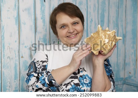 Old woman holding a seashell. Selective focus on her face. Blue marine background. Woman in her sixties.