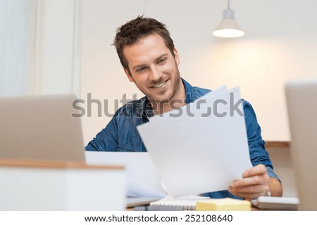 Happy young businessman reading paperwork at desk in office   Royalty-Free Stock Photo #252108640