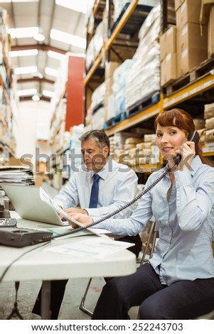 Manager working on laptop and talking on phone at desk in a large warehouse