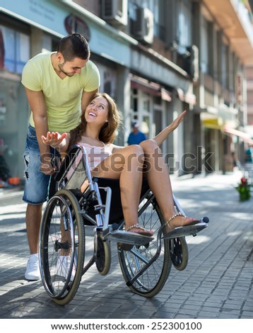 Young caregiver and female in invalid chair laughing at street 