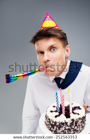 Funny close up picture of young office manager with party hat and blower. Man holding cake and looking at camera
