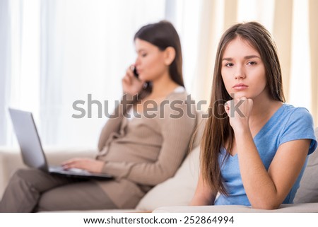 Sad teen girl is sitting on sofa and looking at the camera while her mother is working with laptop.