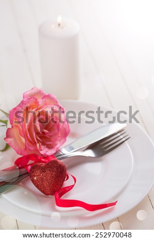 Romantic table setting. White plates, fresh rose, candle  and little red heart.  Selective focus is on heart. 