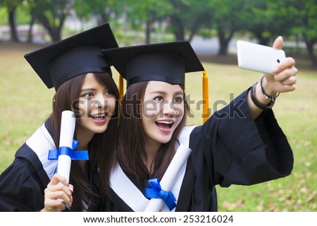 Two happy women in graduation gowns taking picture with cell phone