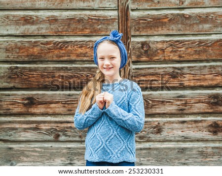 Outdoor portrait of a cute fashion little girl, wearing warm blue pullover and scarf, standing against brown wooden door