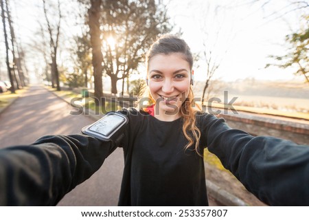 Young Sporty Woman Taking a Selfie at Park. She is Looking at Camera, that is the POV, Point of View, of the photo. She has a Smart Phone Holder on her Arm and Listen Music with Earphones.