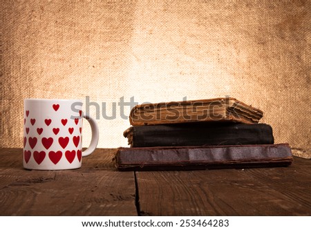 White mug with many pictured hearts and stack of old books on the old wooden table on burlap background.