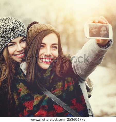 Closeup of two beautiful Caucasian teenage girls with knit beanie hats taking a selfie with smartphone outdoors in winter. Square format images, retouched, instagram look, filter applied.