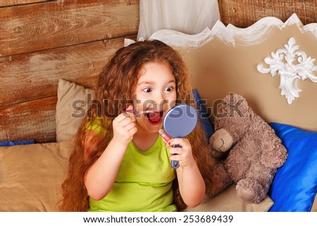 Little girl with long curly hair holds in hands mirror and lipgloss make-up and grimaces