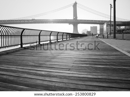 A black and white photograph of the Manhattan Bridge, taken from a wooden walking path in Brooklyn Bridge Park.