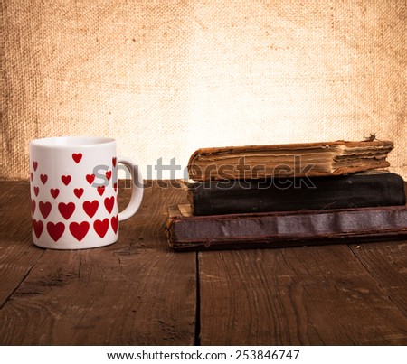 White mug with many pictured hearts and stack of old books on the old wooden table on burlap background.